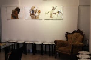 Ściana z grafikami we wnętrzu galerii. Pod trzema obrazami stoi ciąg siedmiu czarnych taboretów z białymi siedziskami oraz fotel uszak,.