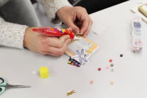 Zbliżenie na dłonie uczestniczki pracującej nad projektem. Osoba nakłada klej introligatorski na białe podobrazie z naklejonym fragmentem tekstu z książki oraz grafiką ważki z perłowym środkiem. Na stole leżą luźne koraliki z literami oraz organizer z zestawami alfabetu.