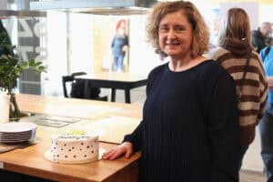 Portret Katarzyny Herman-Gładysz. Uśmiechnięta kobieta o blond, kręconych włosach stoi przy drewnianym blacie na którym leży tort.