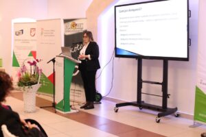 Kobieta w biznesowym stroju prezentuje slajd z definicją i wypunktowanymi rodzajami dostępności (architektoniczna, informacyjno-komunikacyjna, cyfrowa). Baner Ośrodka Wsparcia i Testów oraz kosz z ciemnoróżowymi orchideami stoją obok mównicy