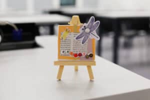 Gotowa praca plastyczna wyeksponowana na małej sztaludze. Przedstawia kompozycję z fragmentem drukowanego tekstu w pomarańczowej ramce. Główną dekorację stanowi grafika fioletowej ważki z perłowym środkiem oraz imię „KRYSTYNA” ułożone z małych koralików-literek u dołu pracy.