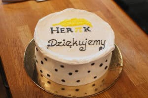 Na drewnianym blacie leży biały, okrągły tort w czarne kropki na złotej podkładce. Na górze napis „HERπK Dziękujemy” i żółty element dekoracyjny