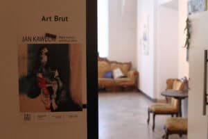 Widok od wejścia do galerii. Na ścianie plakat wystawy Jana Kaweckiego „Najtrwalsza kolekcja Jana”, w głębi jasna sala z meblami