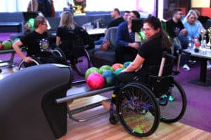 Kobieta w okularach i czarnej koszulce siedzi na wózku inwalidzkim w strefie oczekiwania kręgielni i uśmiecha się, patrzy przez ramię. Obok niej na podajniku leżą kolorowe kule do kręgli. W tle przy stolikach i na kanapach siedzi grupa osób, w tym osoby na wózkach.