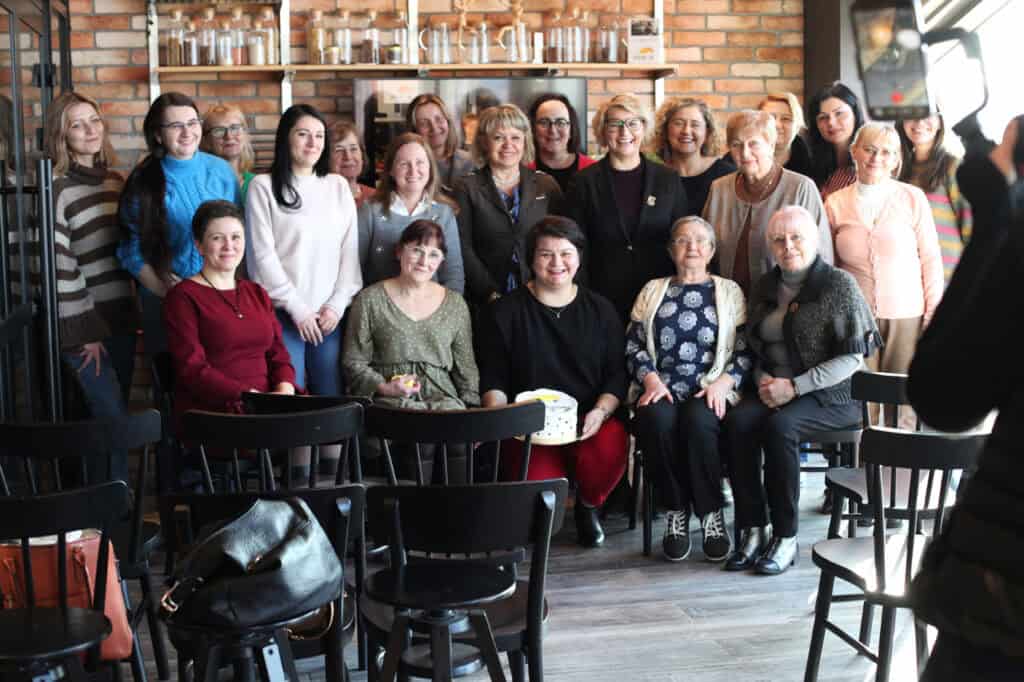 Pamiątkowe zdjęcie grupy około 20 kobiet przed ceglaną ścianą. W centrum siedzi kobieta w czarnej bluzce i czerwonych spodniach, trzyma tort