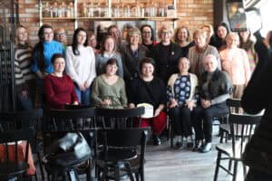 Pamiątkowe zdjęcie grupy około 20 kobiet przed ceglaną ścianą. W centrum siedzi kobieta w czarnej bluzce i czerwonych spodniach, trzyma tort