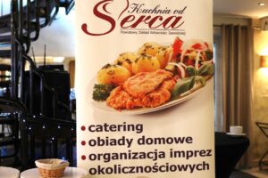 Pionowy baner reklamowy "Kuchnia od Serca" Powiatowego Zakładu Aktywności Zawodowej. Głównym elementem jest zdjęcie obiadu (kotlet, ziemniaki, surówka) oraz wypunktowane usługi: catering, obiady domowe, organizacja imprez okolicznościowych