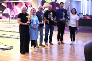 Sześć osób stoi w rzędzie na drewnianej podłodze kręgielni. Część z nich trzyma złote puchary. Po prawej stronie stoi Magdalena Szpyt w białym T-shircie i macha ręką. W tle widoczne są tory do kręgli oraz fioletowo-białe, geometryczne ściany.