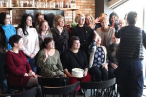 Grupa uczestniczek macha i uśmiecha się do obiektywu. Jedna z kobiet trzyma tort, obok widać osobę nagrywającą smartfonem.