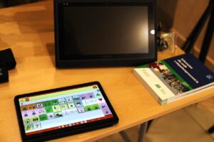 Na drewnianym blacie leżą tablet z aktywną aplikacją do komunikacji alternatywnej (wyświetlającą piktogramy) oraz stos Informatorów ZUS dla osób z niepełnosprawnością 2025