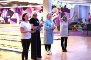 Cztery kobiety stoją przy torze do kręgli. W tle fioletowo-białe ściany. Pierwsza od lewej - Magdalena Szpyt, trzyma notatnik i się uśmiecha. Pozostałe kobiety trzymają złote puchary. Ostatnia trzyma puchar i podnosi ręce w geście triumfu.