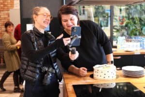 Dwie kobiety stoją obok blatu z białym tortem. Jedna z nich ma na szyi aparat fotograficzny i pokazuje drugiej ekran telefonu umocowanego na stabilizatorze