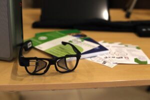 Specjalistyczne okulary z nietypową, siatkową budową (w kolorze czarnym), leżą na jasnym biurku na tle stosu broszur informacyjnych w barwach zieleni i bieli