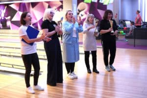 Pięć kobiet stoi w rzędzie przy torach do kręgli. Kobieta po lewej, w białej koszulce (Magdalena Szpyt), klaszcze i trzyma niebieską teczkę. Trzy kobiety trzymają złote puchary. Wszystkie się uśmiechają. W tle widać tory i fioletowo-białe ściany.