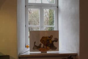 Szeroki, biały parapet okienny w grubym murze, na którym wyeksponowano obraz. Praca przedstawia surrealistyczną, brązowo-żółtą formę przypominającą zniekształconą twarz lub czaszkę na jasnym tle. Przez okno, w tle widać rozmyte drzewa.