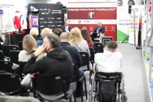 Ujęcie z tyłu na publiczność siedzącą w rzędach krzeseł podczas panelu dyskusyjnego. W prawym dolnym rogu widoczny jest mężczyzna siedzący na wózku inwalidzkim. W tle siedzą dwaj paneliści, a za nimi czerwono-czarny baner „Integracyjne Centrum Sportu i Rehabilitacji 'Start'".