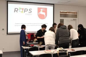Grupa osób stoi przy stole w jasnej sali wykładowej, pracując wspólnie nad materiałami warsztatowymi. W tle widać duży ekran projekcyjny z logo ROPS Lublin oraz herbem województwa lubelskiego. Na ekranie widnieje napis dotyczący finansowania zadania publicznego ze środków PFRON.