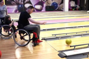 Mężczyzna na czarnym wózku, ubrany w czarną koszulkę i szare spodnie dresowe oraz czerwone buty sportowe, rzuca zielonkawo-żółtą kulę na drewniany tor do kręgli. Kula toczy się w prawo. Mężczyzna śledzi jej tor lotu, mając wyciągniętą prawą rękę. Po lewej, na drugim planie widoczna jest kobieta w okularach siedząca na wózku. W tle widoczne są równoległe, jasne tory, a na ścianie za nimi dominuje duża, fioletowo-biała grafika z motywem kręgli i kul.