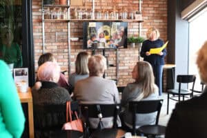 Katarzyna Herman-Gładysz stoi przy ekranie i czyta z żółtej teczki. Przed nią siedzą słuchaczki, w tle ceglana ściana i półki z akcesoriami. Widok z widowni