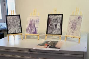 Cztery oprawione grafiki na drewnianych sztalugach w galerii. Przed nimi leży plakat wystawy „ART BRUT” z nazwiskiem artysty.