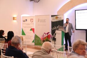 Mężczyzna w szarej marynarce stoi przy mównicy i przemawia, gestykuluje. W tle stoją banery: Ośrodek Wsparcia i Testów, PFRON oraz Ośrodek Szkolno-Wychowawczy. Widok z widowni