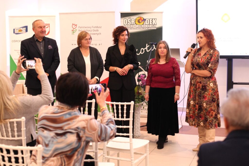 Grupa pięciu prelegentów (jeden mężczyzna, cztery kobiety, m.in. Iwona Nowak-Podkowa, Barbara Sadowska-Burlita, Małgorzata Zarzycka) stoją razem przed publicznością siedzącą na białych krzesłach, tyłem do obiektywu. W tle trzy banery (m.in.: Ośrodek Szkolno Wychowawczy, PFRON). Kobieta w kwiecistej sukience, stojąca z prawej, mówi do mikrofonu, a dwie osoby z publiczności robią zdjęcia smartfonami