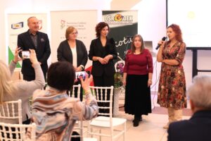 Grupa pięciu prelegentów (jeden mężczyzna, cztery kobiety, m.in. Iwona Nowak-Podkowa, Barbara Sadowska-Burlita, Małgorzata Zarzycka) stoją razem przed publicznością siedzącą na białych krzesłach, tyłem do obiektywu. W tle trzy banery (m.in.: Ośrodek Szkolno Wychowawczy, PFRON). Kobieta w kwiecistej sukience, stojąca z prawej, mówi do mikrofonu, a dwie osoby z publiczności robią zdjęcia smartfonami