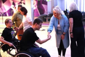 Wnętrze kręgielni. Kobieta w jasnoniebieskim kardiganie podaje rękę mężczyźnie na wózku inwalidzkim w czarnym T-shircie i dżinsach. Obok siedzi drugi mężczyzna na wózku i patrzy w ich stronę. W tle inni uczestnicy i geometryczne dekoracje.