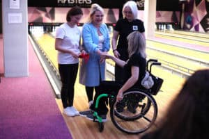 Wręczenie nagrody przy torze do kręgli. Trzy kobiety stoją obok siebie i podają dłoń kobiecie na wózku inwalidzkim. Kobieta na wózku ma jasne włosy i czarny strój sportowy. Jedna z kobiet trzyma małe czerwone opakowanie. W tle tor i logo „MASTERS BOWLING”.