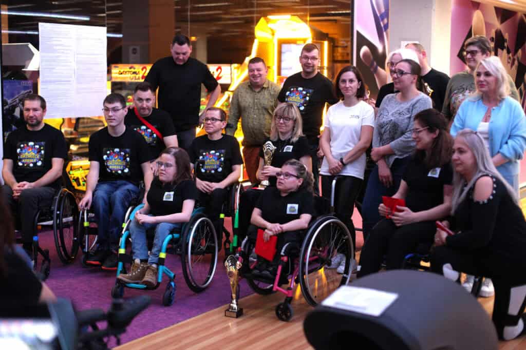 Zdjęcie grupowe w kręgielni. Duża grupa uczestników pozuje do zdjęcia. Wiele osób siedzi na wózkach inwalidzkich. Osoby z przodu trzymają złote puchary i mają czarne koszulki z kolorowym logo. Z tyłu stoją pozostali uczestnicy. Na pierwszym planie stoi złoty puchar.