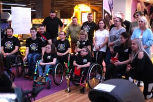 Zdjęcie grupowe w kręgielni. Duża grupa uczestników pozuje do zdjęcia. Wiele osób siedzi na wózkach inwalidzkich. Osoby z przodu trzymają złote puchary i mają czarne koszulki z kolorowym logo. Z tyłu stoją pozostali uczestnicy. Na pierwszym planie stoi złoty puchar.