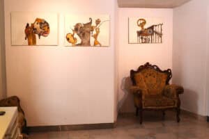 Narożnik sali wystawowej. Na ścianie po lewej wiszą dwie grafiki, a na ścianie po prawej – jedna. W pracach dominują odcienie brązu, żółci i czerni, ukazując fantastyczne postacie i twarze o nieregularnych kształtach. Poniżej obrazów, w samym rogu, stoi dekoracyjny fotel.