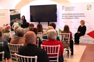 Prelegentka mówi do publiczności. Po prawej stronie sceny siedzi tłumaczka języka migowego ubrana w czarny strój. W tle stoi baner promujący Centra Informacyjno-Doradcze PFRON dla osób z niepełnosprawnością. Widok z widowni.