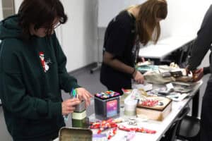 Zbliżenie na uczestników warsztatów pracujących przy stole zastawionym przyborami plastycznymi. Na pierwszym planie osoba w ciemnozielonej bluzie z kapturem wybiera kolorowe mazaki z metalowego pudełka. Na stole leżą zestawy profesjonalnych markerów, pędzle, ozdobne pudełka i inne materiały do rękodzieła. W tle widać drugą osobę pochyloną nad swoimi materiałami.