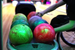 Zbliżenie na stojak z ułożonymi w dwóch pionowych rzędach różnokolorowymi kulami do kręgli, w tym jaskrawozielonej, czerwonej, niebiesko-zielonej i fioletowej, z których część jest lekko podświetlona na zielono, tworząc metaliczną poświatę. Po prawej stronie widoczna jest wyciągnięta ręka osoby na wózku, sięgająca po jedną z kul na dalszym planie.