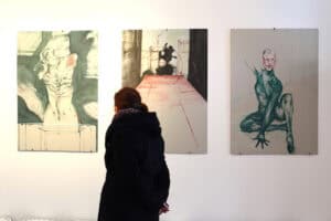 Kobieta ogląda trzy wielkoformatowe obrazy Jana Kaweckiego na białej ścianie galerii.