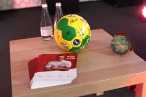 Drewniany stolik kawowy z pamiątkami Michała Szyby: żółto-zieloną piłką ręczną z autografami, brązowym medalem „Qatar 2015” i ulotkami promocyjnymi, w tle dwie szklane butelki wody