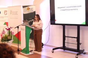 Monika Wac, Dyrektor PFRON Oddział Lubelski wygłasza prezentację. Ekran za nią wyświetla slajd: "Aktywność i uczestnictwo osób z niepełnosprawnością. Perspektywa indywidualna i społeczna". Obok mównicy widać kosz z fioletowymi orchideami i banery PFRON oraz Ośrodka Szkolno-Wychowawczego