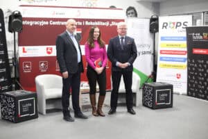 Trzy osoby: dwóch mężczyzn i kobieta w różowym swetrze stoją obok siebie na szarym dywanie podczas wydarzenia prasowego. W tle widoczny jest duży czerwono-czarny baner promujący „Integracyjne Centrum Sportu i Rehabilitacji 'Start'" oraz baner ROPS z hasłem „Wszyscy jesteśmy tacy sami", a także baner "Lublin, Miasto Inspiracji."