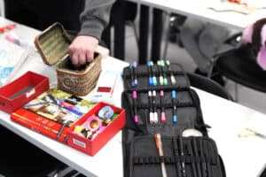 Zbliżenie na przybory artystyczne rozłożone na białym stole. Widoczny jest otwarty czarny piórnik z kolorowymi cienkopisami, czerwone pudełko z rolkami ozdobnych taśm, zestaw kredek oraz pleciona skrzyneczka, do której sięga dłoń uczestnika. W tle widać fragmenty materiałów plastycznych na sąsiednim stanowisku.