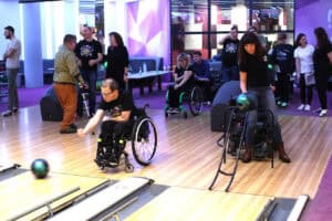 Wnętrze kręgielni i strefy wypoczynku, z kilkoma osobami grającymi w kręgle lub oczekującymi na swoją kolej. Na pierwszym planie, mężczyzna w okularach na wózku inwalidzkim rzuca ciemnozieloną kulę na tor. Po prawej, kobieta w czarnej koszulce i dżinsach korzysta z rampy, aby wprowadzić kulę na tor. W tle stoi i siedzi na kanapach grupa ludzi, w tym kilka osób na wózkach. W tle fioletowe ściany i nowoczesne oświetlenie.