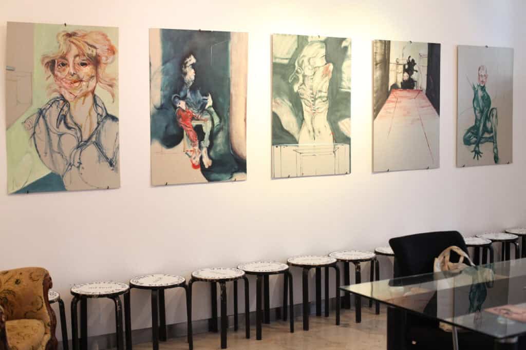 Wnętrze galerii z pięcioma obrazami na białej ścianie. Pod nimi stoją czarne taborety z napisem „TEATROTERAPIA LUBELSKA"