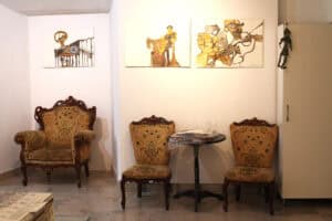 Wystawa z trzema grafikami na białych ścianach. Poniżej nich, w narożniku, stoi duży fotel uszak, a obok niego, przy mniejszym fragmencie ściany, dwa pasujące do niego mniejsze krzesła ustawione po obu stronach okrągłego, ciemnego stolika. Na stoliku leżą przedmioty zabezpieczone folią. Po prawej stronie widać fragment białej szafy, na której zawieszona jest mała, ciemna figurka pajacyka.