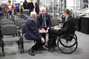 Hala wystawowa z szarym dywanem. Dwóch mężczyzn (w centrum kadru prof. Robert Litwiński) w granatowych marynarkach rozmawia z elegancko ubranym mężczyzną siedzącym na wózku inwalidzkim (Piotr Jacniacki). Wokół stoją puste, szare krzesła ustawione w rzędy. W tle, po prawej stronie, widać gabloty z eksponatami sportowymi.