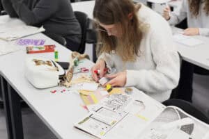 Młoda kobieta w białym, puszystym swetrze i okularach, pracuje przy stole, skupia się na detalach swojego projektu. Skleja małe, drewniane elementy, używając kleju w tubce. Przed nią leżą wycinki z gazet, kolorowe papiery, ozdobne naklejki i biała torebka z aplikacją misia. W tle widać inne osoby pracujące w skupieniu.