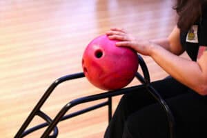 Różowa kula do kręgli leżąca na metalowym stojaku. Z prawej strony widoczna jest kobieta (częściowo poza kadrem), która delikatnie trzyma dwiema rękami wierzch kuli, przygotowując ją do rzutu. Kobieta ubrana jest w czarną koszulkę z logo „GRZMIĄCE KRĘGLE". Tło stanowi rozmyty, jasny, drewniany tor do kręgli.