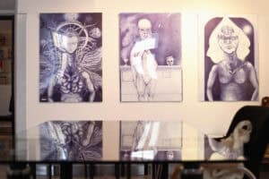 Trzy wielkoformatowe grafiki w odcieniach fioletu i błękitu wiszą na białej ścianie. Na pierwszym planie widać szklany stół z odbiciem prac