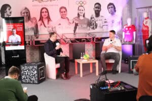 Michał Szyba i prowadzący siedzą w jasnych fotelach przy stoliku z piłką i medalem, w tle wystawa „Historia sportem pisana” z wizerunkami sportowców i gablotami z trofeami