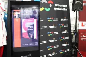 Od lewej duży, pionowy monitor dotykowy (INTBOARD) wyświetla plakat informujący o premierze książki pt. „Integracyjne Centrum Sportu i Rehabilitacji 'Start' w Lublinie. Historia i teraźniejszość (1945-2025)" autorstwa Roberta Litwińskiego. Obok monitora stoi duży, czarny baner z powtarzającym się napisem „Centrum Historii Sportu w Lublinie" i logotypem MOSiR Lublin.