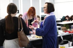 Prowadząca w niebieskim kardiganie (dr Anna Bieganowska-Skóra) rozmawia z uczestniczkami warsztatów, trzymając w dłoniach małe drewniane listewki. Na stole między nimi leżą materiały do dekoracji i naklejki. W tle widać inne osoby pracujące przy swoich stanowiskach w jasnej sali.